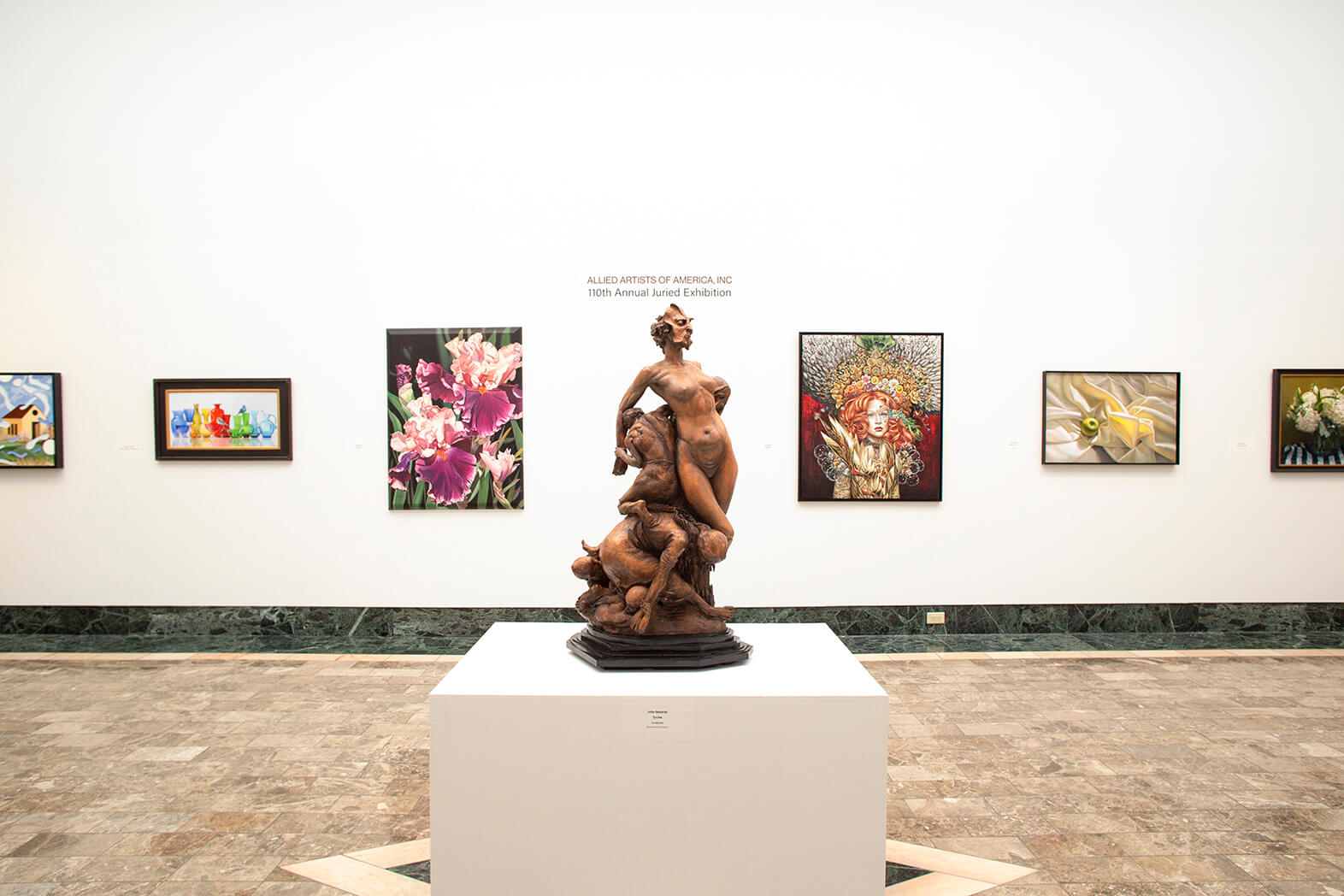 A gallery displays multiple paintings along a white wall and in the center on a pedestal is a sculpture of multiple people on a wooden base.