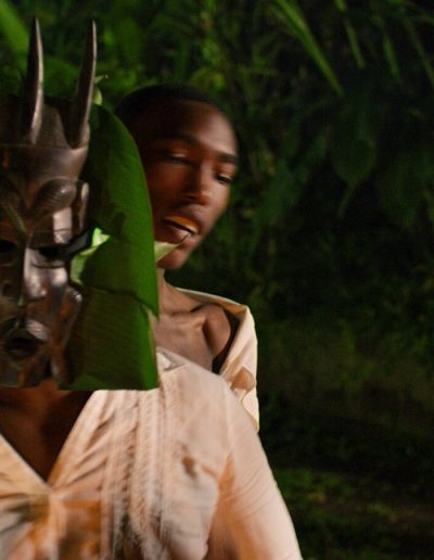 A black man standing behind another black man who is wearing a ceremonial mask and white blouse surrounded by leaves and trees