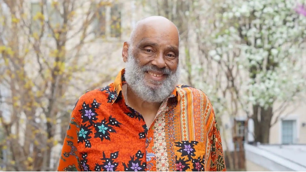 A photo portrait of a man from the chest up standing outdoors. The man is partially bald with a brown complexion and white-grey facial hair. He smiles and looks at the camera, he is wearing a brightly colored button up collared shirt with a mixed pattern in orange, yellow, black blue and purple,