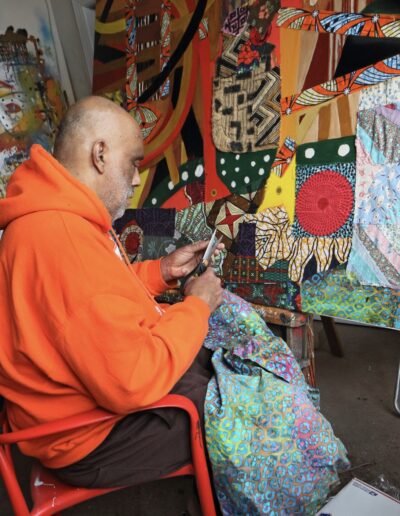 An artist is an orange hoodie sits in a chair, using scissors to cut a piece of vibrant, multi colored patterned fabric. Behind him is a large, complex collage or textile artwork featuring a mix of geometric shapes, bold colors, and various fabric swatches